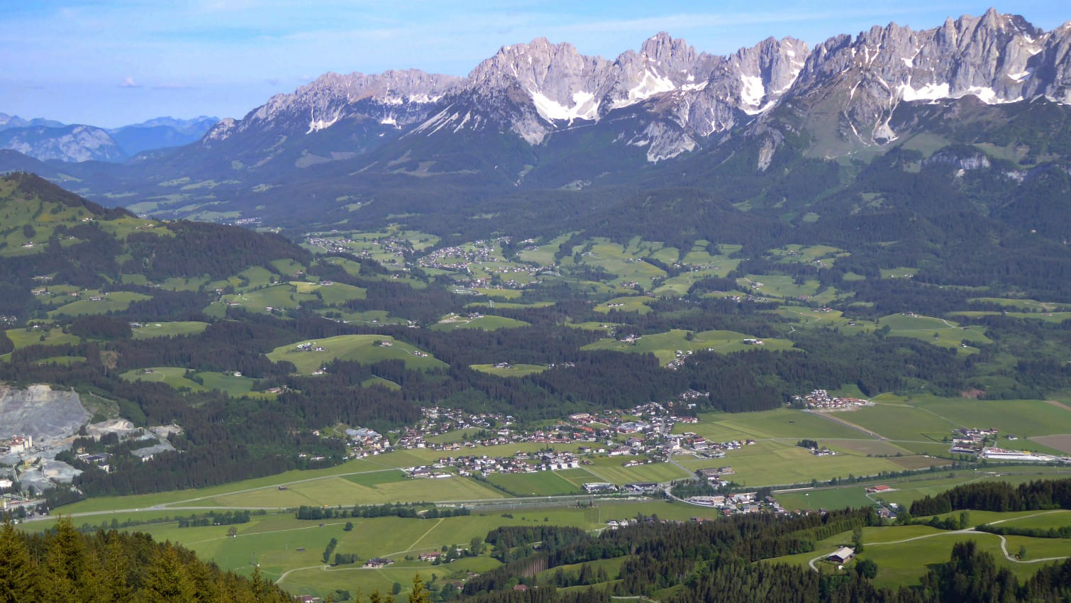 Die Aufnahme zeigt eine weite Landschaft mit grünen Wiesen, Wäldern und verstreuten Häusern in einem Tal, in dem die Gemeinde Oberndorf liegt. Im Hintergrund erhebt sich die Gebirgskette Wilder Kaiser mit schneebedeckten Gipfeln unter einem klaren blauen Himmel.