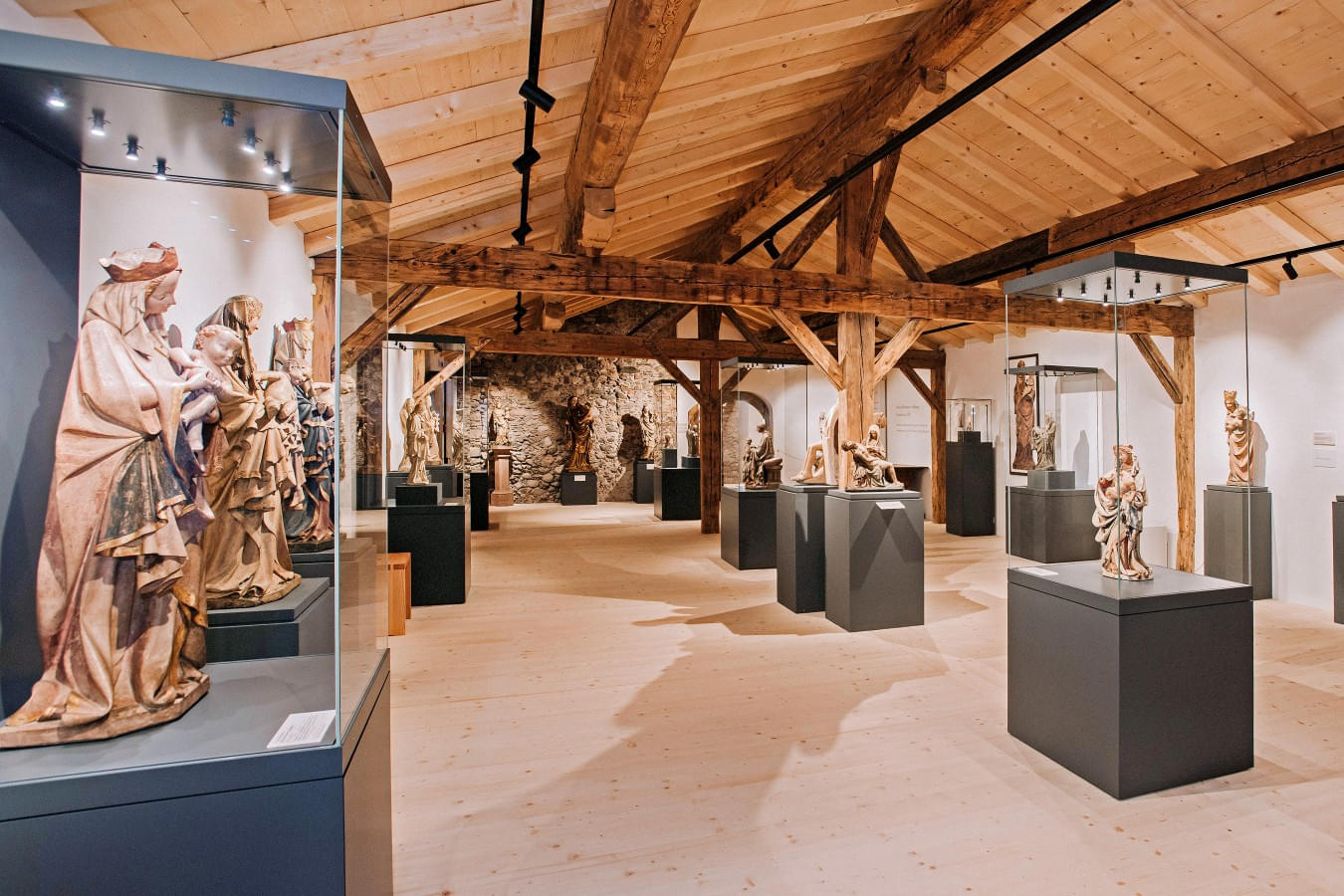 Ausstellungsraum mit Holzdecke und -balken, der mittelalterliche Skulpturen zeigt, die auf Podesten und in Vitrinen präsentiert werden.